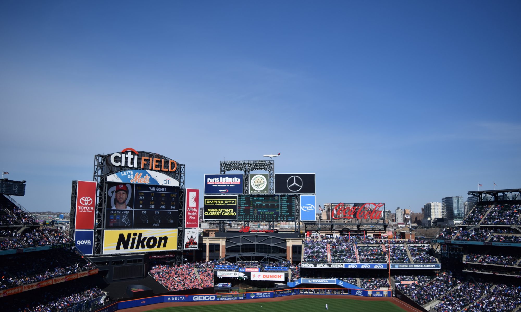 photo of Citi Field from the first base promenade, spanning from the LF stands to the RF stands, with a plane flying over head